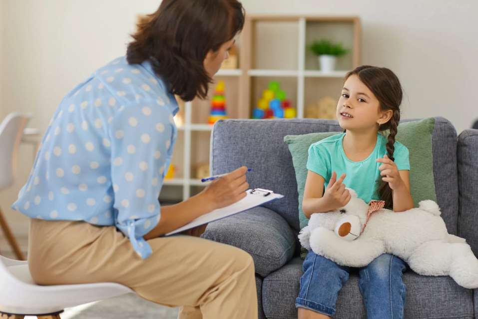 An image of a girl sitting on a sofa holding a teddy bear talking to a woman on a chair opposite holding a clipboard